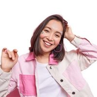 Horizontal shot of optimistic carefree Asian woman tilts head smiles sincerely dressed in fashionable jacket being in good mood isolated over pink background. Happy emotions and feelings concept
